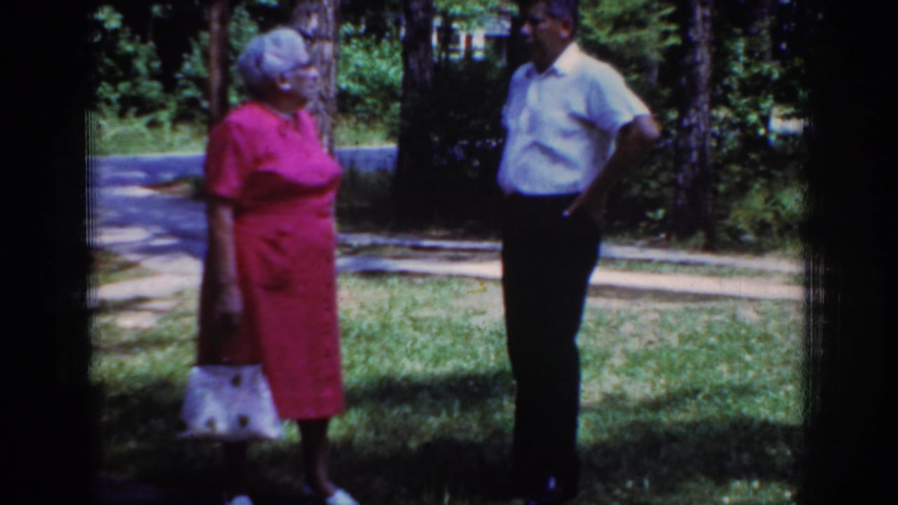 In Raleigh home-movie footage from 1962, an older woman with a white handbag stands facing an older man in a shaded yard.