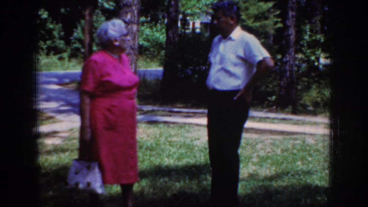 Older woman and man stand apart on grass near trees and a paved drive.