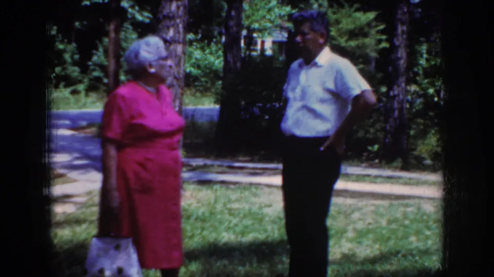 A closer 8mm frame shows the woman in pink holding a white handbag as the man listens in the wooded yard.