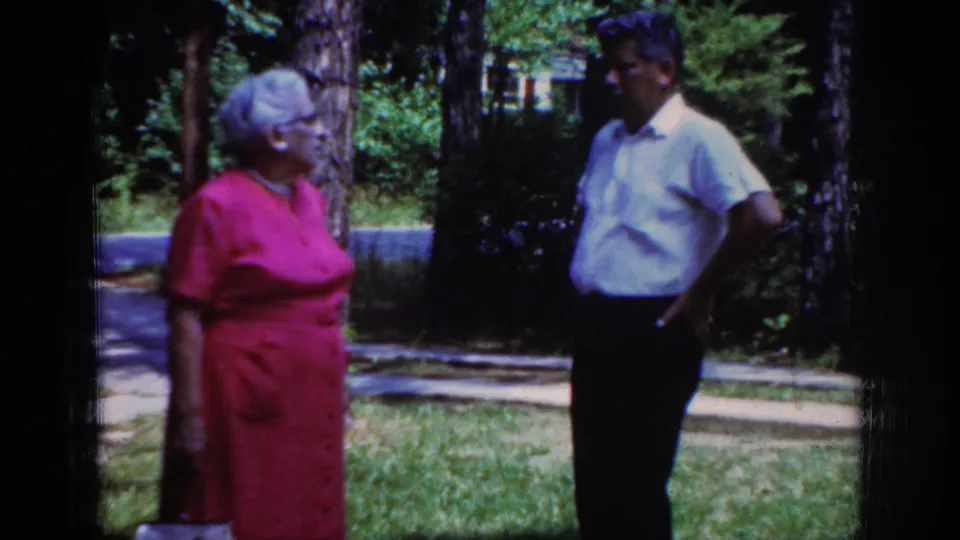 The 1962 Raleigh yard scene continues with the older woman turned toward the man under trees and patchy sunlight.