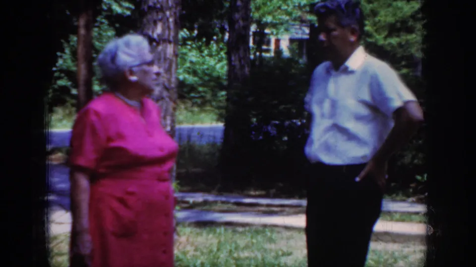 A home-movie frame from Raleigh in 1962 shows the older woman in profile and the man facing her on a sunny residential lawn.