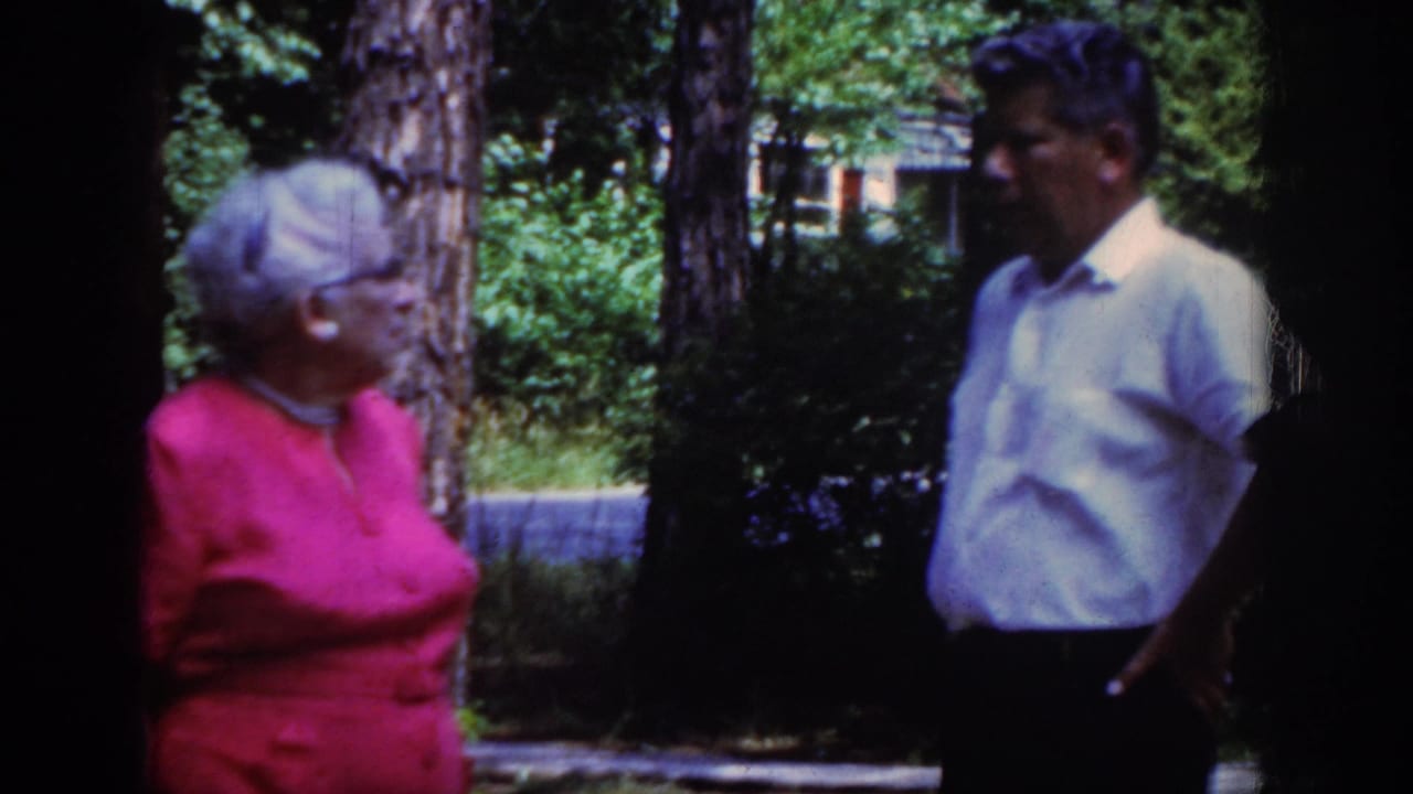 1962 Raleigh home-movie frame of an older woman in a red dress and glasses speaking with a man in a white short-sleeved shirt outdoors.