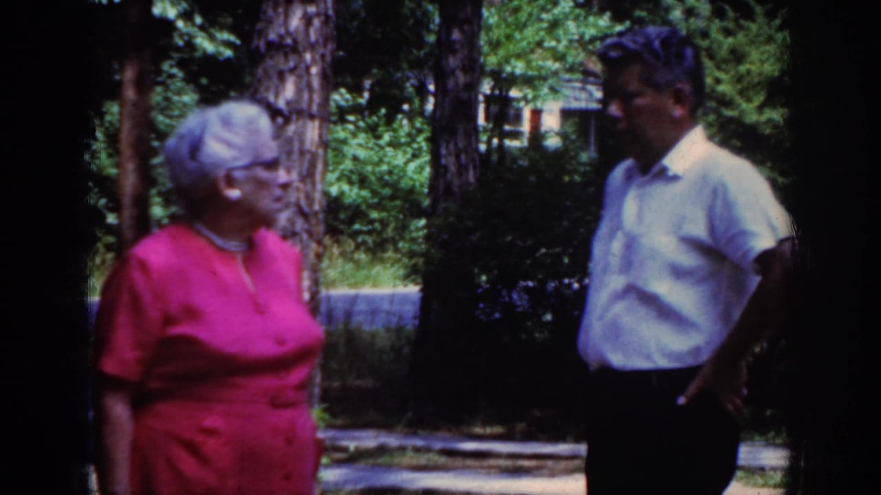 Woman in red and man in white stand apart while talking near trees and shrubs.