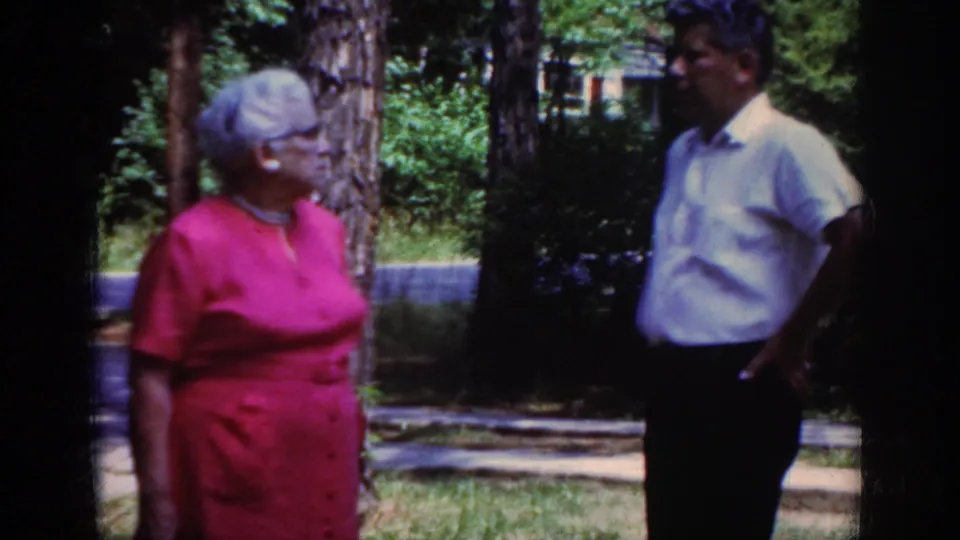 Home-movie frame from Raleigh in 1962 showing an older woman in red looking toward a man in a white shirt in a shaded residential yard.