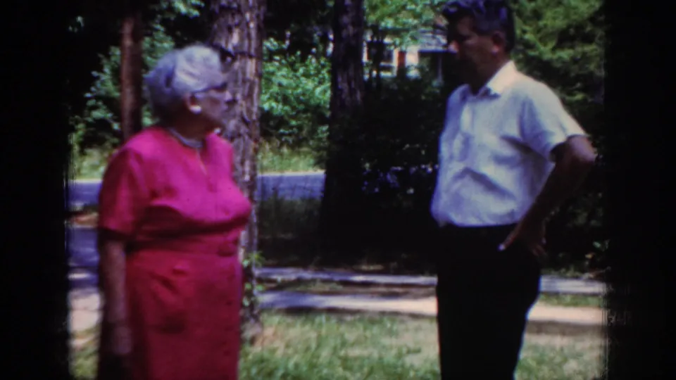 1962 Raleigh home-movie frame of a man in a white short-sleeved shirt facing an older woman in red amid trees and lawn.
