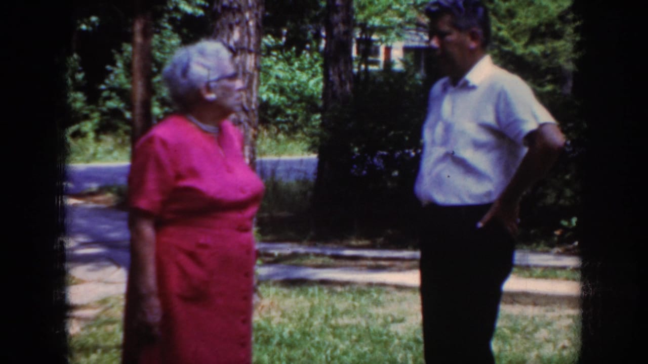 In metadata-supported Raleigh, 1962, two adults converse in a sunlit yard with trees, grass, and a path behind them.