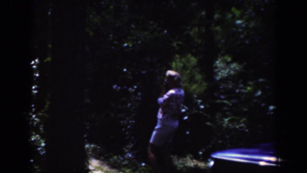 A woman in a patterned top and light shorts stands in a wooded outdoor area, with part of a parked car visible at right.
