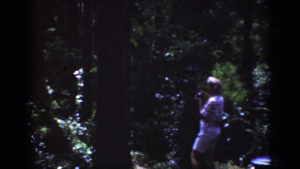 A woman stands in a dark wooded area with dappled sunlight, holding her hands near her chest while facing left.