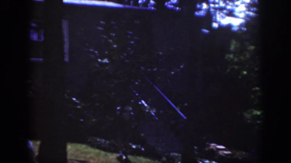 A dim 1962 8mm frame shows the hiker partly lost in forest shadow, with blue-green highlights and a narrow diagonal light streak.