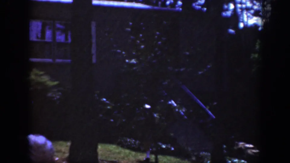 1962 Raleigh home-movie frame showing a dark residential yard, house windows, trees, shrubs, and a partial person at the lower edge.