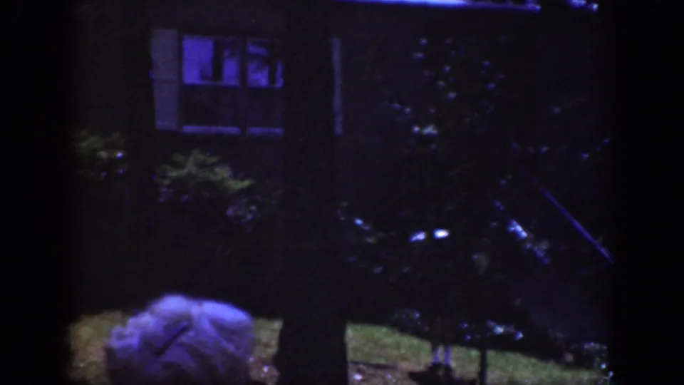 1962 Raleigh nighttime exterior with house windows behind trees and a light-colored hat entering the lower part of the frame.