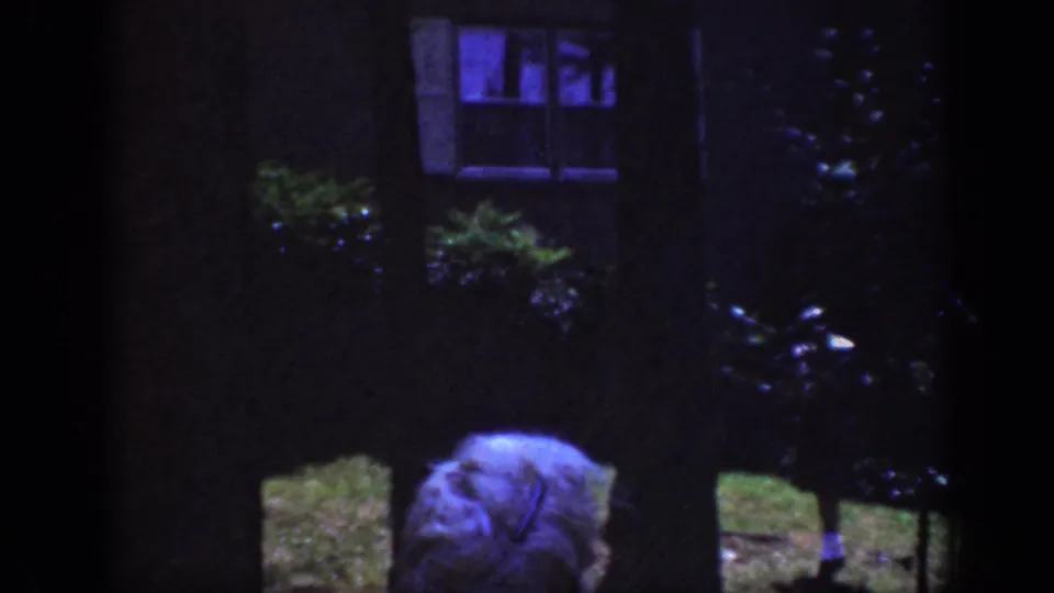 1962 Raleigh home-movie frame with a light-colored hat passing through a dark yard beneath trees near a house window.