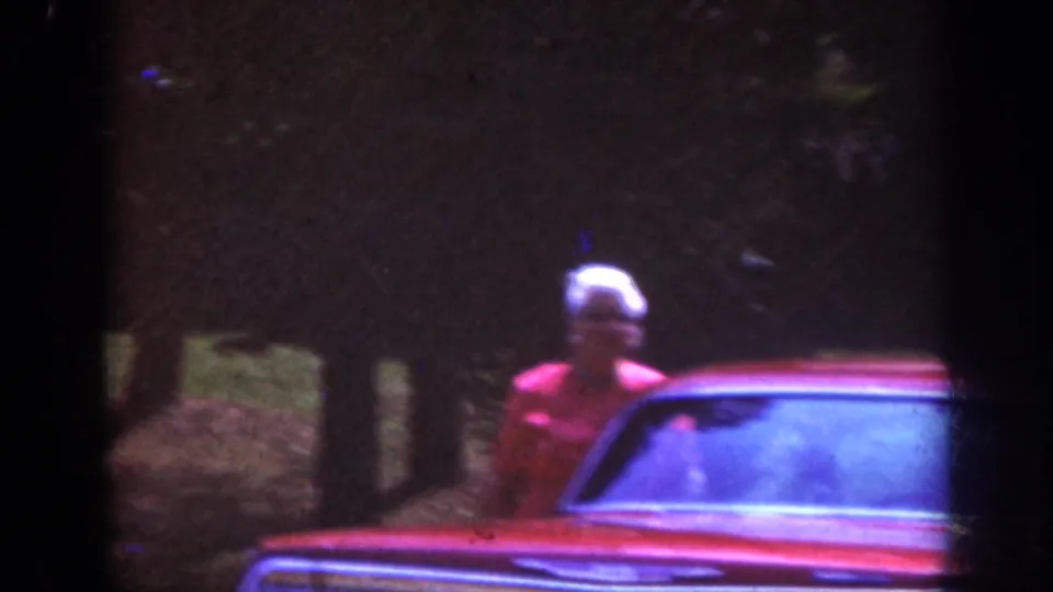 1962 Raleigh home-movie frame showing a person in a light hat near a red car during a nighttime arrival outside a home.