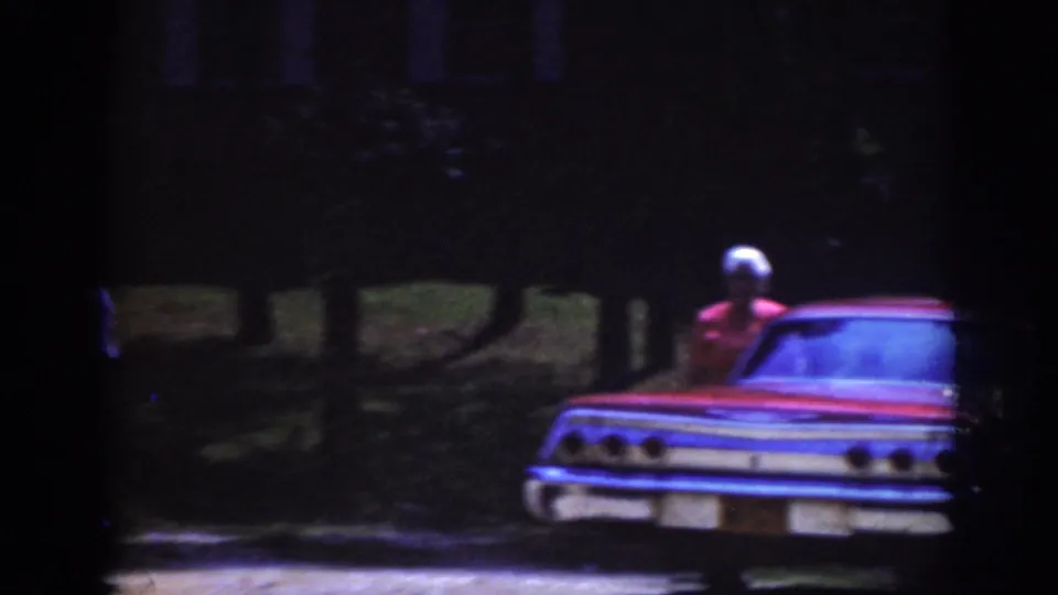 1962 Raleigh 8mm frame showing the rear of a red older-model car with an older woman nearby in darkness.