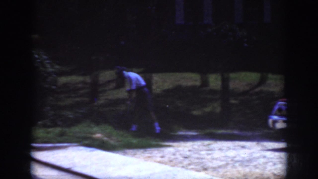 1962 Raleigh home-movie frame showing a distant person tending a grassy area near trees, with curb pavement and a partial car visible.
