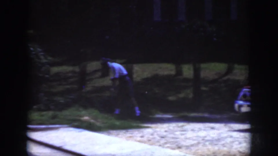 1962 Raleigh home-movie frame showing a distant person tending a grassy area near trees, with curb pavement and a partial car visible.