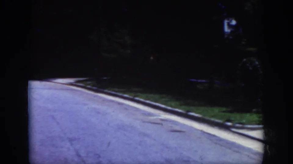 1962 Raleigh home-movie frame showing a quiet paved street or driveway, curb, shaded grass, and trees with no clear person centered.
