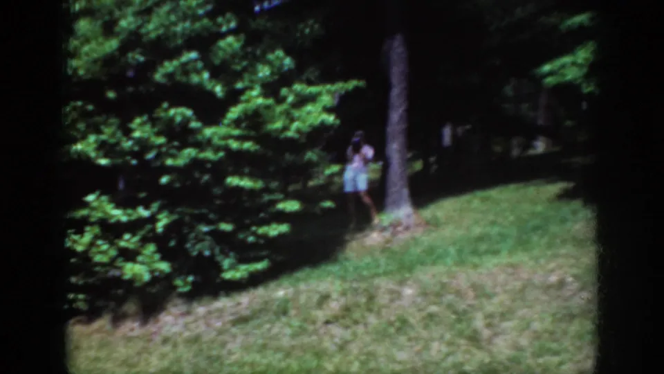 1962 Raleigh home-movie frame showing a distant person near a tree trunk, shrubs, and a sloped residential lawn in bright green foliage.