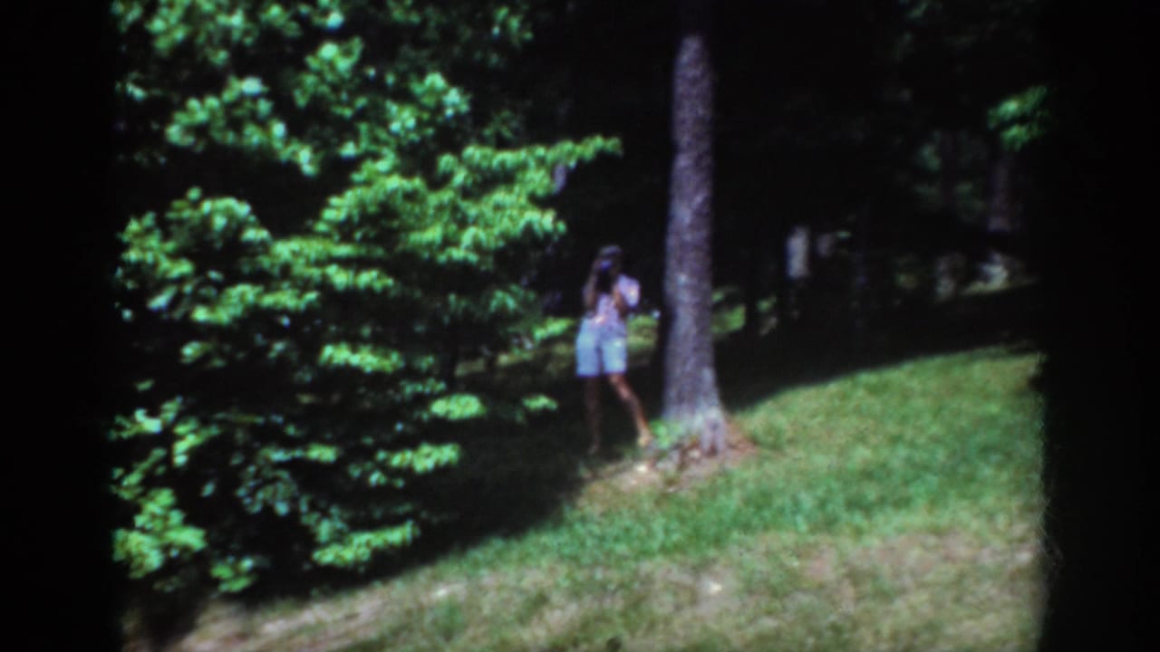 1962 Raleigh home-movie frame showing a woman in shorts holding a camera beside a tree in a sunlit wooded park.