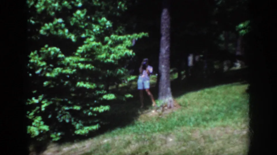 1962 Raleigh home-movie frame showing a woman in shorts holding a camera beside a tree in a sunlit wooded park.