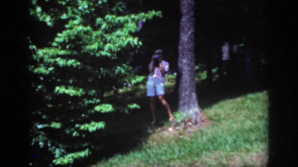 1962 Raleigh 8mm frame of a woman filming near a tree, surrounded by green foliage and strong sunlight.