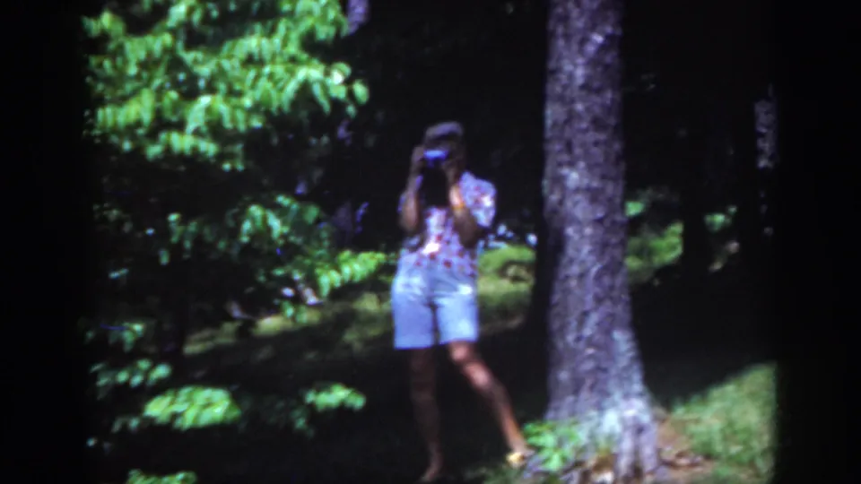 1962 Raleigh home-movie still of a woman filming in a wooded park, partly shaded by dense green foliage.