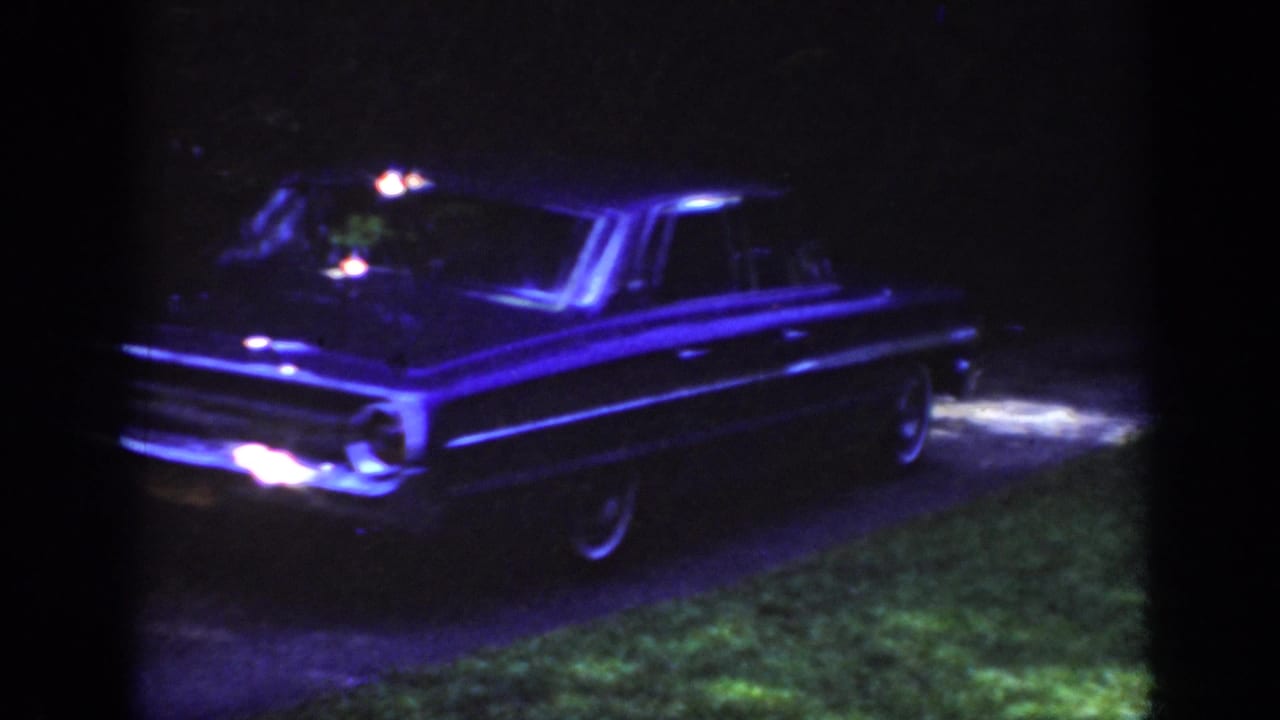 Classic sedan with headlights on along a dark driveway beside grass.