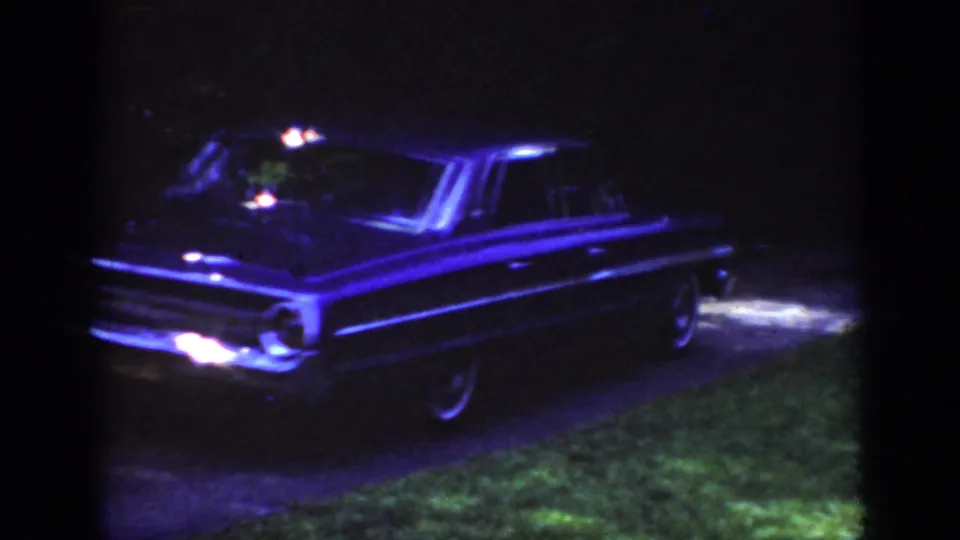 Raleigh 1962 frame of a classic American sedan lit blue at night on a driveway near lawn and trees.