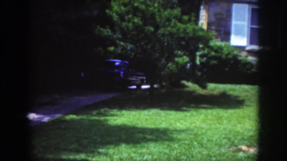 Raleigh 1962 frame showing a dark car near shrubbery and a visible house window beside a nighttime lawn.