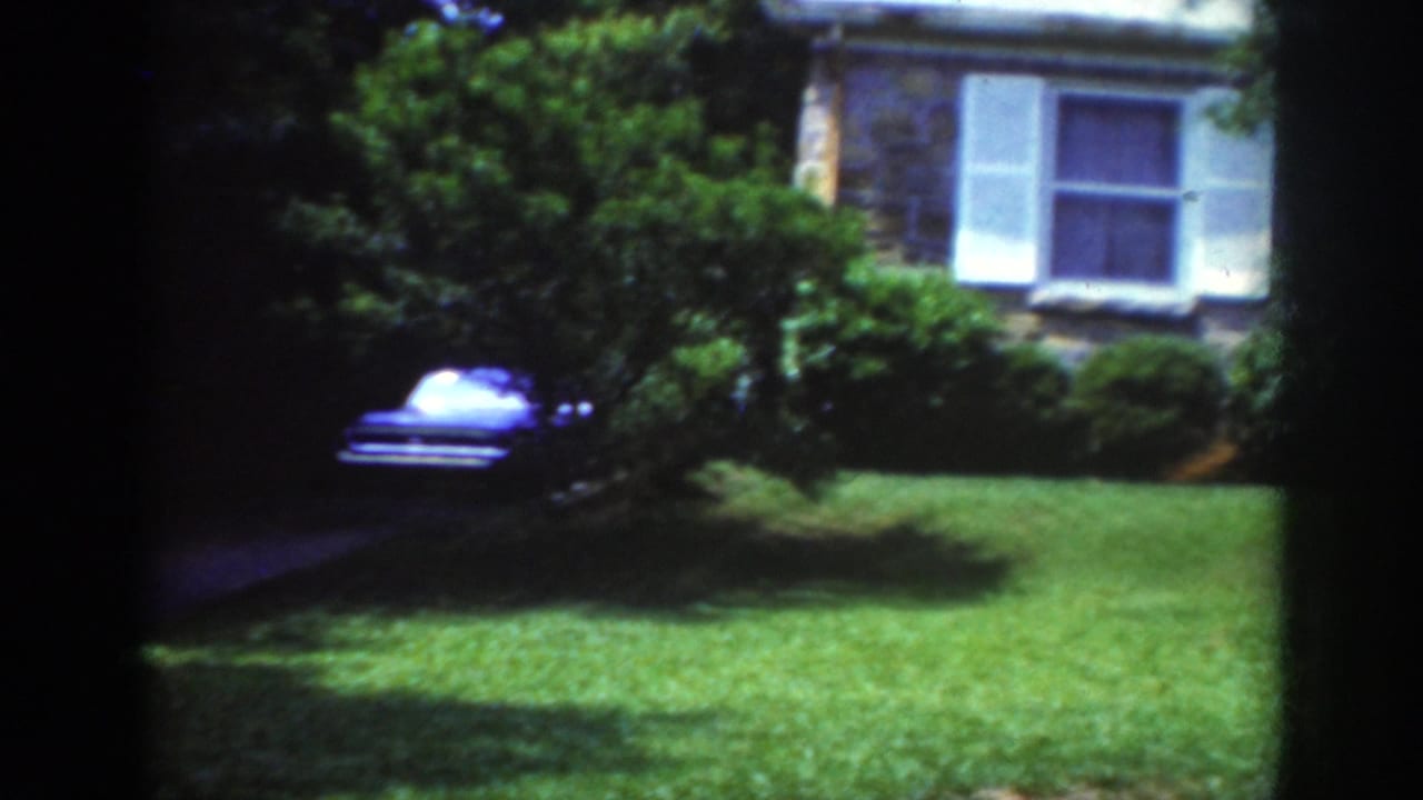 1962 Raleigh home-movie frame showing a stone residential exterior with white shutters, lawn, trees, and a blue car near the driveway.