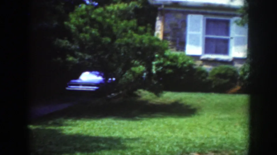 1962 Raleigh home-movie frame showing a stone residential exterior with white shutters, lawn, trees, and a blue car near the driveway.