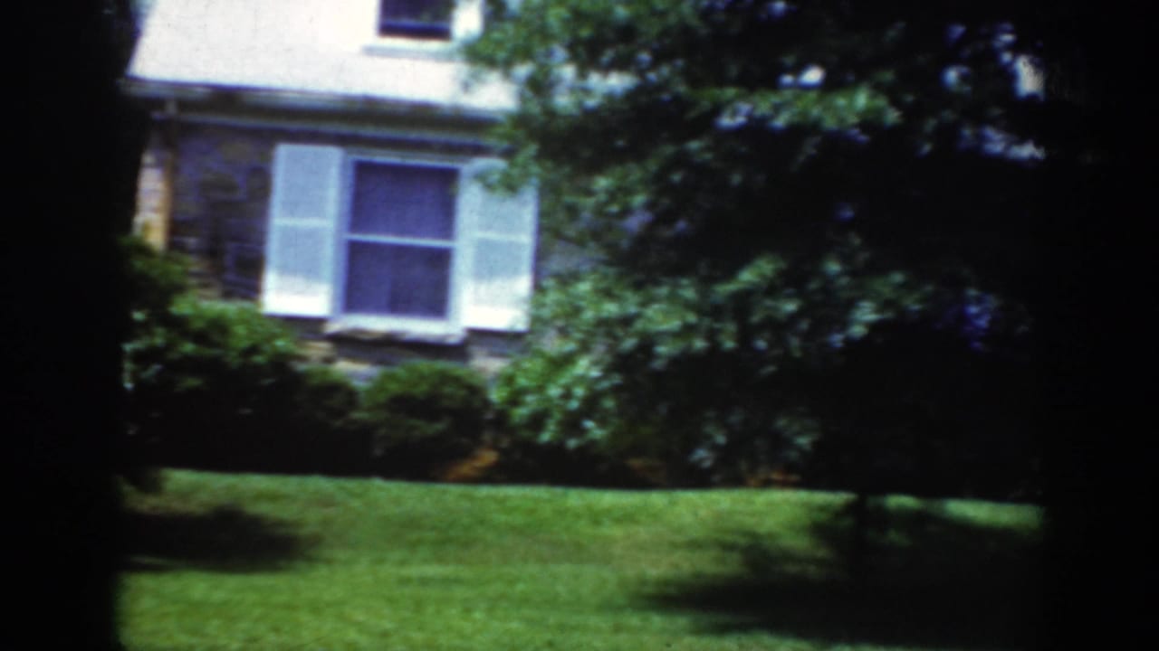 1962 Raleigh frame showing a residential stone facade, white shutters, trimmed shrubs, green lawn, and a dark vertical frame edge.