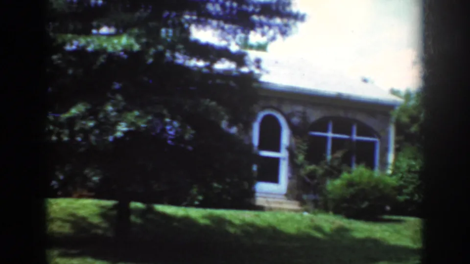 1962 Raleigh home-movie view of a stone house entrance with an arched door, large window, bushes, and a shaded lawn.