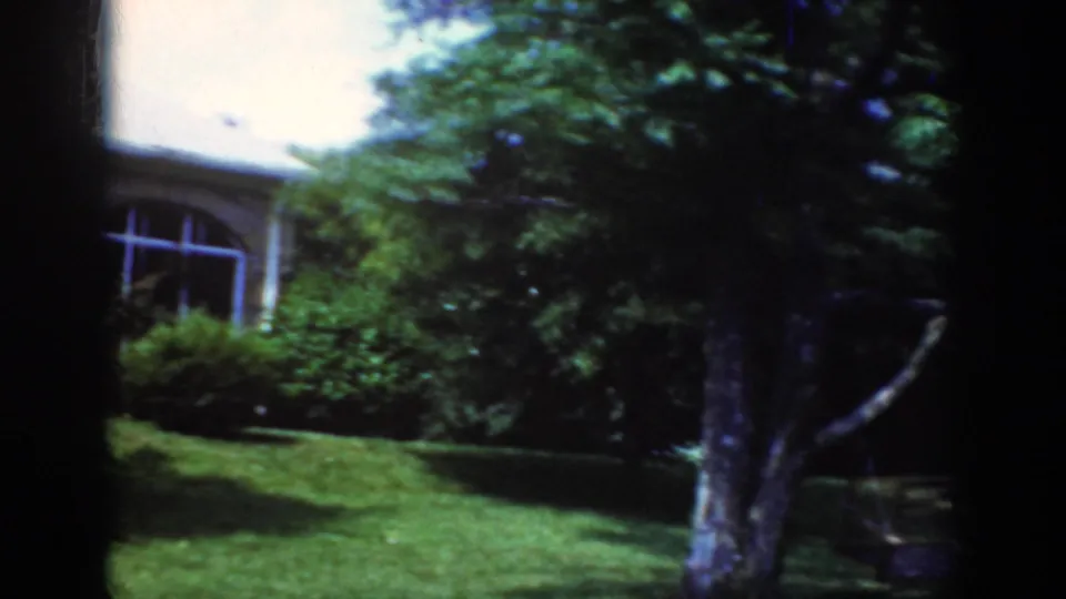 1962 Raleigh frame showing a leafy residential exterior with a stone house partly hidden by a tree, shrubs, and green lawn.