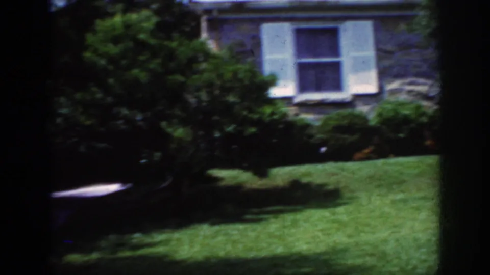 1962 Raleigh frame showing a house window with white shutters, stone exterior, shrubs, and a sunlit lawn.