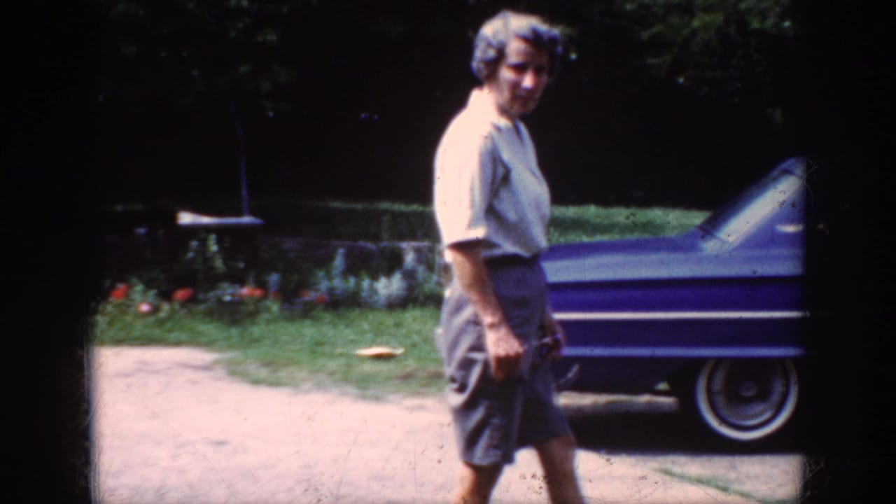In Raleigh, North Carolina, 1962, an older woman in a light shirt and gray shorts walks past a blue sedan in a residential yard.