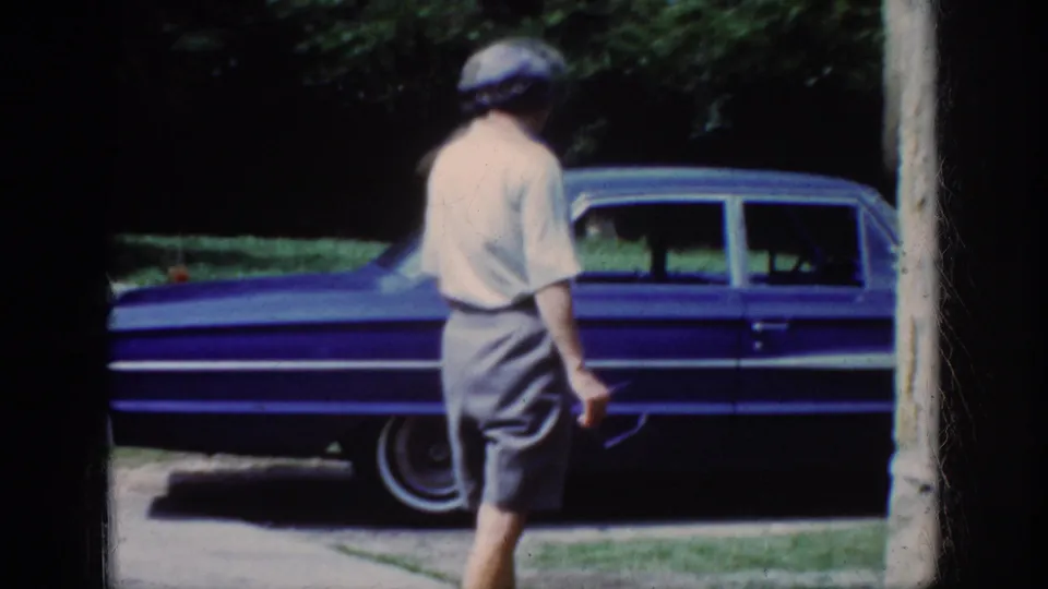 The woman is viewed from behind as she walks near the parked blue sedan, with a lawn and wooded background visible.