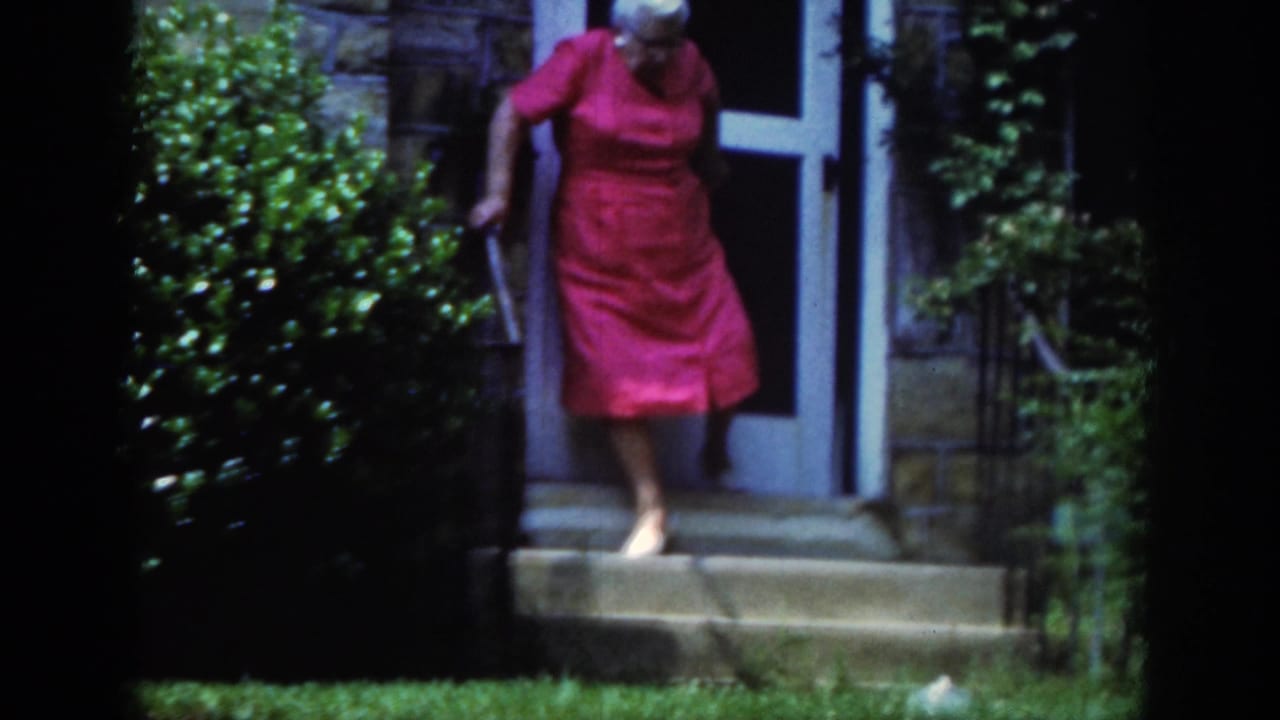 Elderly woman in a red dress steps down from a doorway while holding a cane.