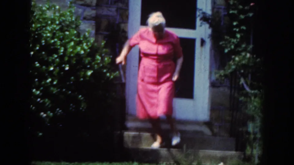 An elderly woman with white hair moves down the steps in front of a stone-faced Raleigh house entrance, framed by greenery.