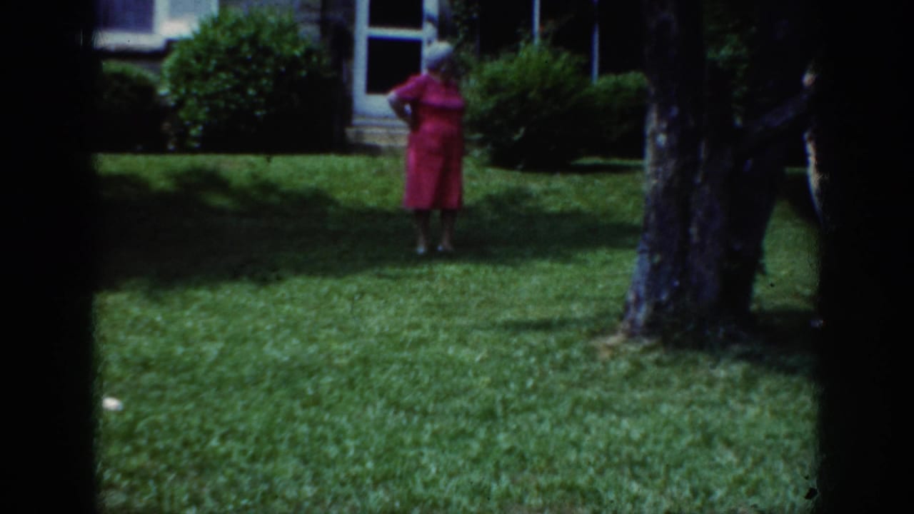 In 1962 Raleigh home-movie footage, a woman in a pink dress and light hat stands on a lawn near a house and shrubs.