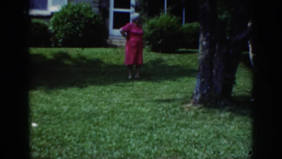 In 1962 Raleigh home-movie footage, a woman in a pink dress and light hat stands on a lawn near a house and shrubs.