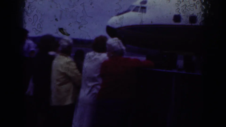 A group of adults stands near the nose of a small airplane on an airfield, with soft 8mm color and visible film marks.
