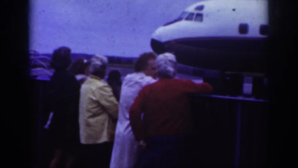 Adults, including a figure in light clothing and another in red, stand near a small airplane at the airfield in Raleigh footage.