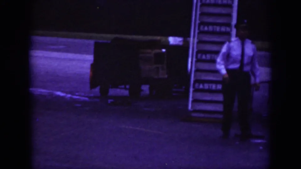 A uniformed airport or airline worker stands beside boarding stairs and baggage carts near an aircraft in Raleigh, 1962.
