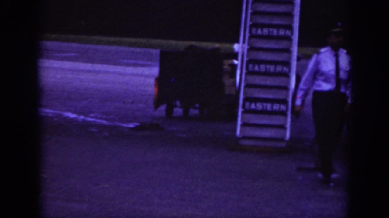 Raleigh 1962 airport ramp scene showing an Eastern Airlines stairway, a worker in a white shirt, and cargo equipment in low light.