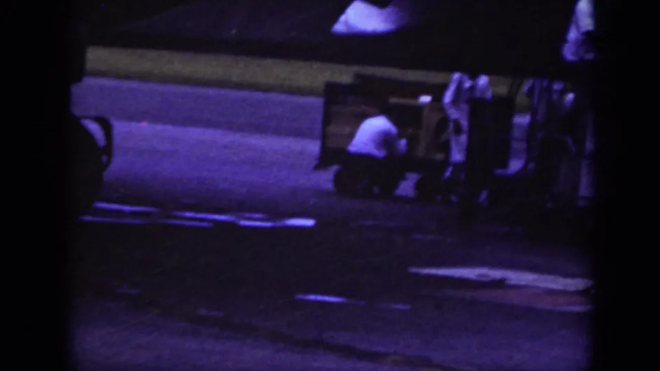 Raleigh 1962 airport ground scene with a worker crouched beside cargo equipment and a baggage cart under low night light.