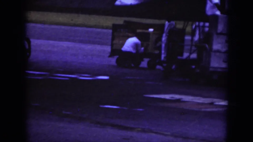 Raleigh 1962 airport ramp view with a ground crew worker crouched near cargo equipment and a baggage cart in purple-tinted film.