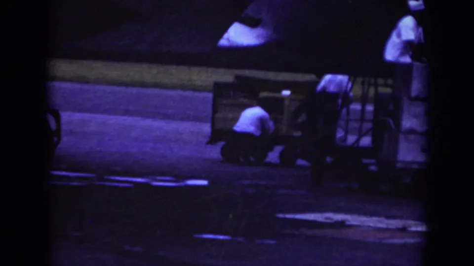 Raleigh 1962 airport ground crew scene with one worker crouched near cargo equipment and another partly visible beside a baggage cart.