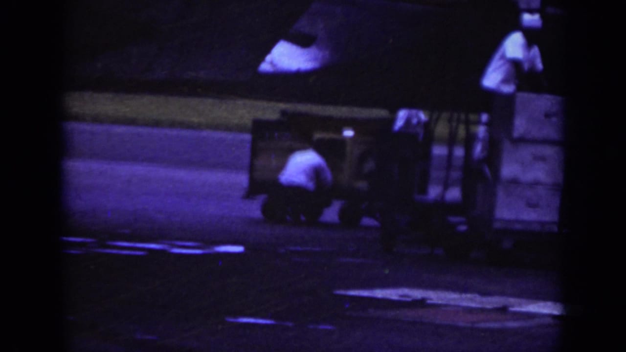 1962 Raleigh airport home-movie frame showing workers near a baggage trailer on the runway under dark, color-shifted exposure.