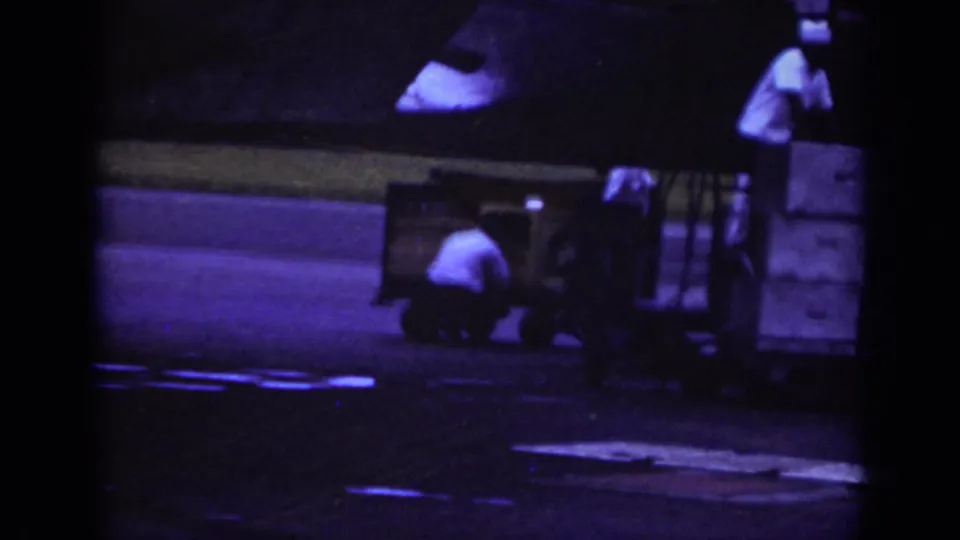 1962 Raleigh airport home-movie frame showing workers near a baggage trailer on the runway under dark, color-shifted exposure.